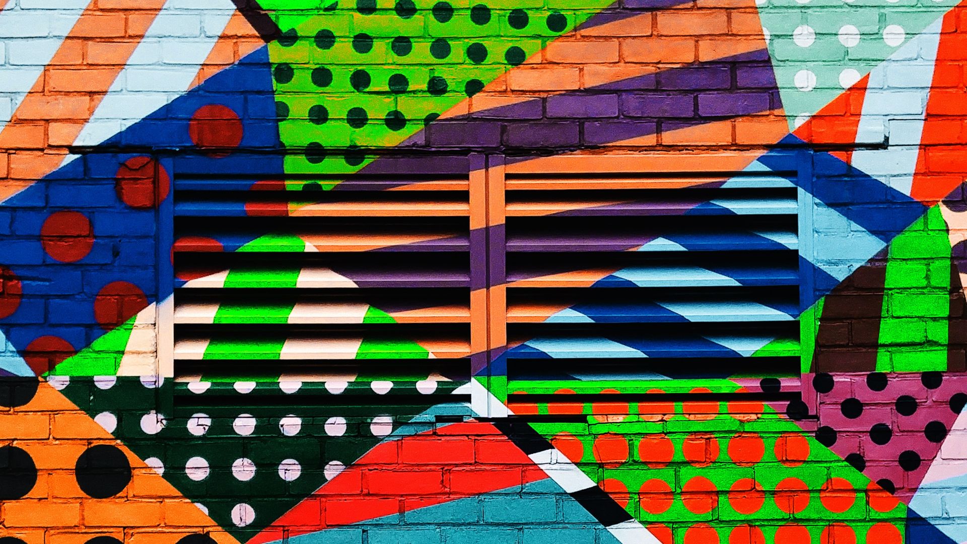 A collage of brightly colored, geometric patterns painted on a brick wall with vents.