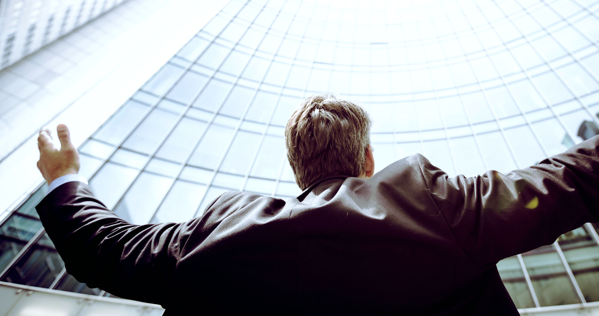 A male corporate employee staring up to a tall corporate building with his arms stretched out.